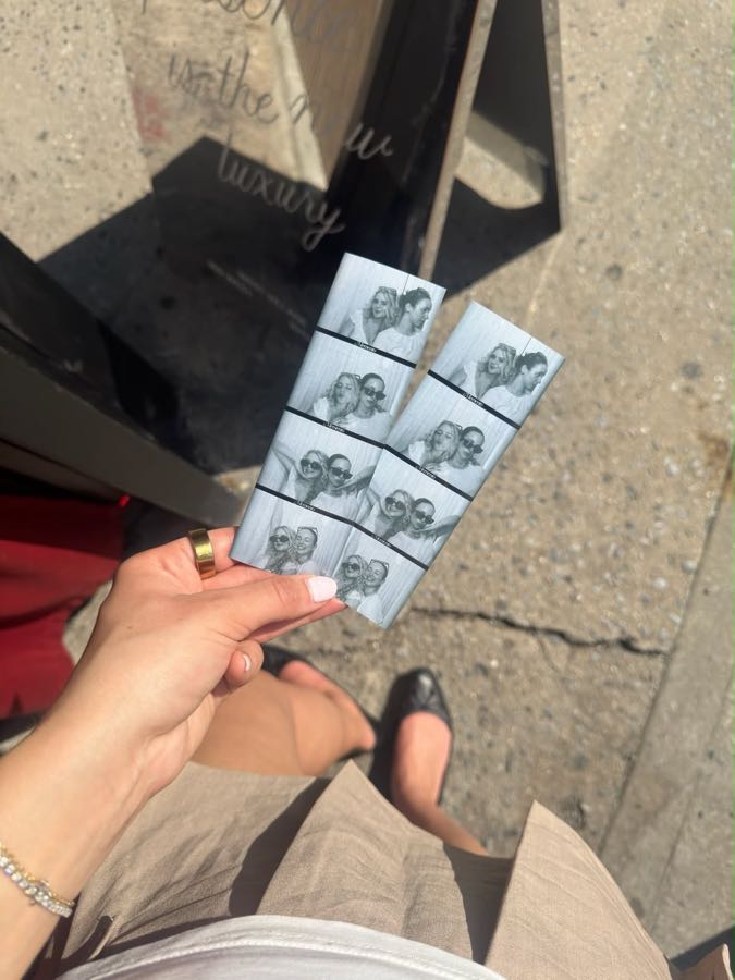 A hand holding two fresh photo strips on a sunny West Village sidewalk.